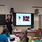 Alcune studentesse delle elementari guardano una professoressa che spiega il sistema immunitario con l'ausilio di una presentazione multimediale.